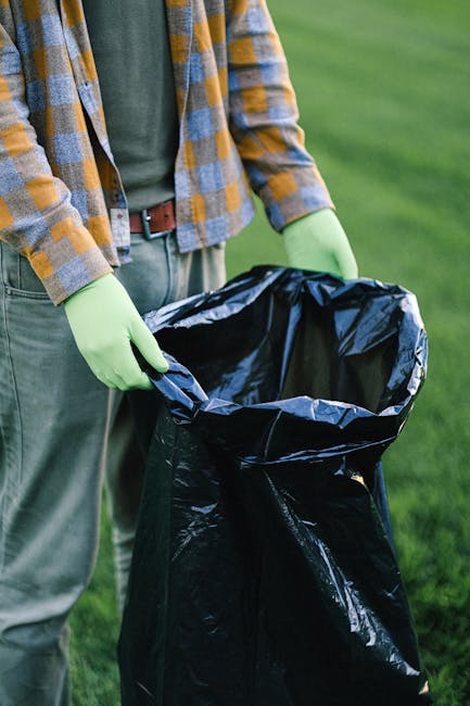 A person dressed in casual outdoor clothing, including a plaid shirt with blue, yellow, and gray tones and gray trousers, is holding a black rubbish bag open with both hands, wearing light green gloves for protection. The bag appears to be made of shiny plastic material, with its top edge slightly crumpled as it is being prepared for rubbish collection. The setting is an outdoor area with green grass visible in the background, suggesting a garden or park environment. The scene is well-lit with natural daylight, emphasizing the cleanliness and professionalism of waste handling. This image illustrates a typical scene of private waste disposal or rubbish collection, which may be associated with alternative rubbish removal services provided by companies like Rubbish Removal Camden, highlighting the importance of on-site clearance and proper waste management practices in residential or outdoor settings.