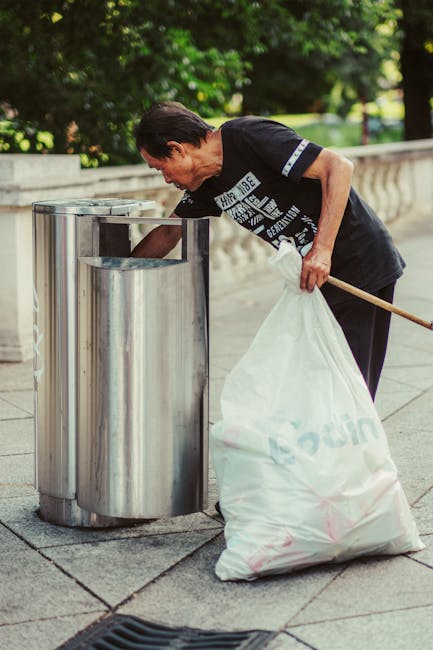 A large pile of mixed waste and rubbish bags is accumulated around a central recycling bin in an outdoor urban setting, situated on a paved area near a steel railing. The recycling bin, labeled for mixed paper and cardboard, has its lid open, overflowing with crumpled paper, flattened cardboard boxes, and other paper waste. Surrounding the bin are various black plastic rubbish bags, some torn open, revealing contents such as discarded packaging and household waste. Several brown cardboard boxes, some flattened and others still intact, are scattered on the ground, along with additional smaller items like plastic wrappers, a beverage carton, and a pink plastic container. Behind the waste collection area, a grey car is parked to the left, and a multi-storey building with scaffolding and blue netting covers the facade, indicating ongoing construction or maintenance. In the background, storefronts with signs and a partially visible red recycling or waste container contribute to the busy urban environment. The scene illustrates an instance of on-site waste accumulation that could relate to private rubbish collection services like those provided by Rubbish Removal Camden, highlighting the importance of proper waste management in urban areas.