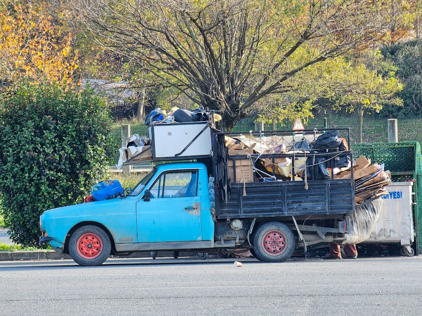 A small, old blue pickup truck with red wheels is parked on the side of a street, loaded with various types of bulky rubbish including cardboard boxes, plastic bags, and miscellaneous debris. The truck's cargo area is filled to capacity with items stacked and leaning, some tied with ropes to secure the load. In the background, there is a large leafy tree with branches extending overhead, and a green metal fence lining the area behind the vehicle. The scene appears to be in a residential or urban area during daylight hours, with natural light illuminating the scene and casting soft shadows. This image visually relates to private rubbish collection or alternative waste handling services, as the vehicle is used for on-site clearance of household or commercial waste, consistent with the services offered by Rubbish Removal Camden.