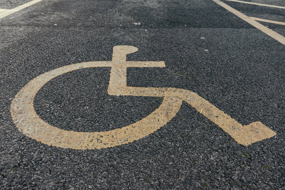 A painted yellow wheelchair accessible symbol on a dark asphalt surface, indicating designated parking or access for disabled individuals. The symbol consists of a circular wheelchair representation with a handlebar and a seat, with an attached diagonal line extending to the right. The asphalt shows a textured, rough surface with small gravel particles visible, and faint lines or markings can be seen in the background. The yellow paint appears weathered and slightly worn, with some chipping and fading, suggesting outdoor exposure. The overall scene is in a parking lot or street environment, with natural daylight illuminating the area, highlighting the contrast between the yellow symbol and the dark asphalt. This image appears relevant to the context of accessible spaces, which is sometimes associated with maintaining clear areas free of obstructions, such as rubbish or clutter, possibly involving private or independent waste removal services provided by companies like Rubbish Removal Camden for accessible and compliant rubbish disposal.