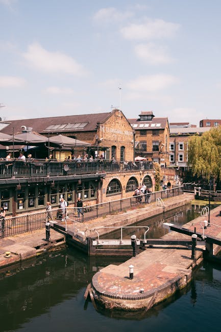 A daytime scene of a canal-side building with a brick and stone facade, featuring arched windows and an outdoor terrace with black metal railings, occupied by several people sitting under umbrellas. The building is situated on the edge of a waterway with calm, dark water, and a small stone pier extends into the canal with metal railings and steps leading down to the water. Surrounding the area, there are additional brick and wood structures, along with a tree with green foliage visible on the right side of the scene. The sky is partly cloudy with soft light illuminating the scene, reflecting a typical urban outdoor setting that could be managed through private rubbish removal services such as those offered by Rubbish Removal Camden, which specializes in waste collection for commercial and residential properties in the area.