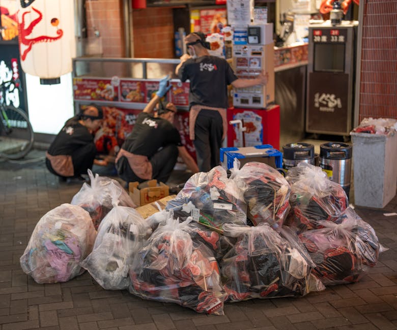 An outdoor urban scene featuring several large transparent plastic bags filled with mixed waste, including clothing and miscellaneous household items, placed on a brick pavement. In the background, three workers dressed in black uniforms with brown aprons and gloves are engaged in sorting or handling additional rubbish near a street-side fast-food outlet or convenience store. The scene is illuminated by ambient lighting, highlighting the textures of the plastic waste bags and the surrounding environment. The workspace includes a small blue toolbox, metal trash bins, and a large red and white food stall with signage and packaging typical of local takeaway shops. This image exemplifies private waste collection or on-site clearance activities, reflecting services offered by companies such as Rubbish Removal Camden involved in the efficient removal and disposal of rubbish in busy urban areas. The overall setting illustrates the process of managing accumulated waste outside commercial premises, suitable for contextual understanding of private rubbish removal services in Camden and similar locations.